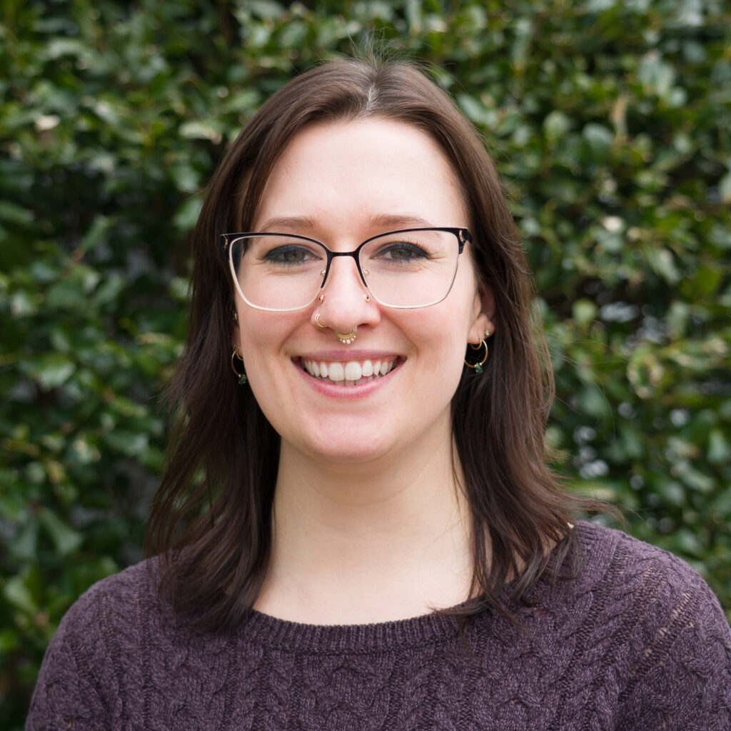 Smiling at the camera, a woman with brown hair and glasses in a brown sweater stands outdoors before green foliage, celebrating Earth's beauty—captured in photo _DSC4787-Edit.