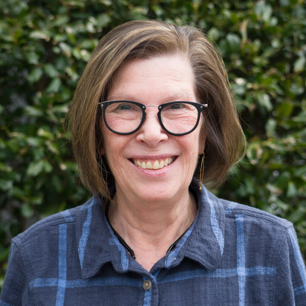 A woman with short brown hair and black glasses smiles at the camera in a blue plaid shirt, surrounded by lush greenery—_DSC4802-Edit captures Earth's natural treasury.