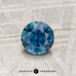 A 0.84-Carat Montana Sapphire (Heated) with a brilliant cut shines blue on a textured light gray surface, with "EARTH'S TREASURY" displayed below.