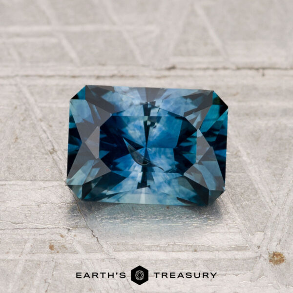 1.65-Carat Montana Sapphire (Heated) A 1.65-carat heated Montana Sapphire, featuring mixed light and dark blue facets, rests on a textured gray surface with the "Earth's Treasury" logo below.