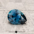 A faceted, pear-shaped blue 1.18-carat Montana sapphire rests on a textured, light-colored surface; "Earth's Treasury" is written below the stone.
