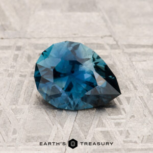 A faceted, pear-shaped blue 1.18-carat Montana sapphire rests on a textured, light-colored surface; "Earth's Treasury" is written below the stone.