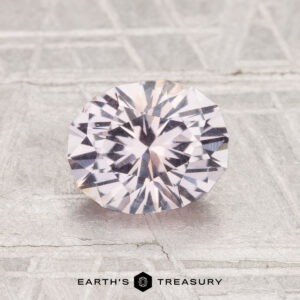 A 1.51-carat round brilliant-cut Montana sapphire with a pale pinkish hue is displayed on a textured, light surface. "EARTH'S TREASURY" is written at the bottom.