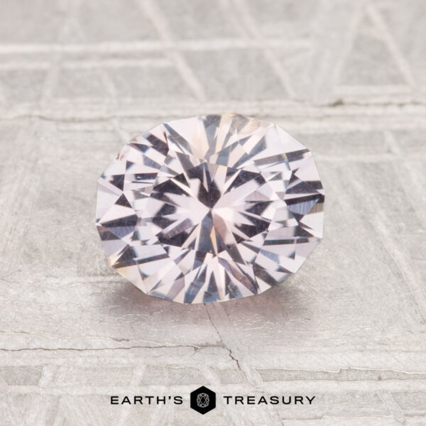 1.51-Carat Montana Sapphire A 1.51-carat round brilliant-cut Montana sapphire with a pale pinkish hue is displayed on a textured, light surface. "EARTH'S TREASURY" is written at the bottom.
