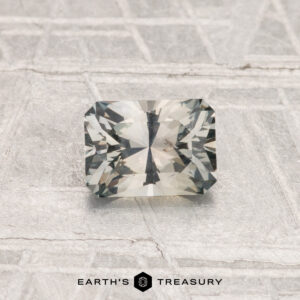 A rectangular, faceted 0.83-carat Montana Sapphire with a light gray hue is displayed on a textured, light-colored surface. The text "EARTH'S TREASURY" appears at the bottom.