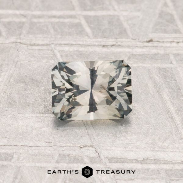 0.83-Carat Montana Sapphire A rectangular, faceted 0.83-carat Montana Sapphire with a light gray hue is displayed on a textured, light-colored surface. The text "EARTH'S TREASURY" appears at the bottom.
