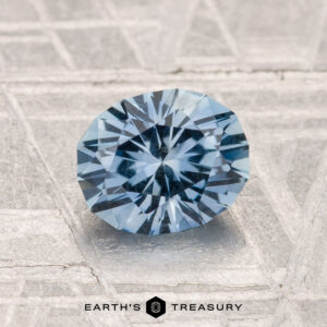 A 1.38-carat Montana sapphire with faceted blue hues is displayed on a textured gray surface, with the Earth's Treasury logo beneath it.