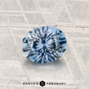 A 1.13-carat Montana sapphire with an oval facet rests on a textured metallic surface, with "Earth's Treasury" inscribed below.