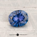A 1.27-carat Montana Sapphire, oval and blue, rests on a textured light surface with the "Earth's Treasury" text and logo displayed below.