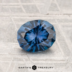 A 1.27-carat, round, faceted Montana sapphire is displayed on a textured gray surface with "EARTH'S TREASURY" at the bottom.