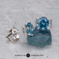 Montana Sapphire Earrings–Teal Blue, 6.0mm (Heated) in silver settings are displayed beside a rough blue stone on a textured surface. "Earth's Treasury" logo appears at the bottom.
