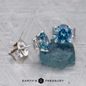 Montana Sapphire Earrings–Teal Blue, 6.0mm (Heated) in silver settings are displayed beside a rough blue stone on a textured surface. "Earth's Treasury" logo appears at the bottom.