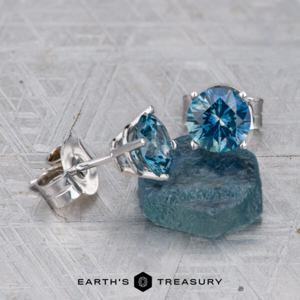 Montana Sapphire Earrings- Teal Blue, 6.0mm (Heated) Montana Sapphire Earrings–Teal Blue, 6.0mm (Heated) in silver settings are displayed beside a rough blue stone on a textured surface. "Earth's Treasury" logo appears at the bottom.