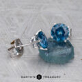A pair of Montana Sapphire Earrings- Peacock Blue, 6.5mm (Heated), in silver settings beside a rough blue sapphire on a textured gray surface. Earth’s Treasury logo appears at the bottom.