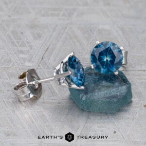 A pair of Montana Sapphire Earrings- Peacock Blue, 6.5mm (Heated), in silver settings beside a rough blue sapphire on a textured gray surface. Earth’s Treasury logo appears at the bottom.