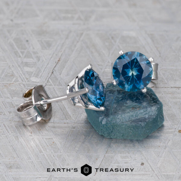 Montana Sapphire Earrings- Peacock Blue, 6.5mm (Heated) A pair of Montana Sapphire Earrings- Peacock Blue, 6.5mm (Heated), in silver settings beside a rough blue sapphire on a textured gray surface. Earth’s Treasury logo appears at the bottom.
