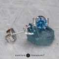 A pair of Montana Sapphire Earrings—teal blue, 5.25mm (heated)—in silver settings, with one earring resting on a rough blue crystal atop a textured gray surface.