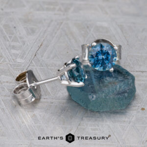 A pair of Montana Sapphire Earrings—teal blue, 5.25mm (heated)—in silver settings, with one earring resting on a rough blue crystal atop a textured gray surface.