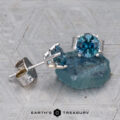 A pair of Montana Sapphire Earrings- Teal, 5.25mm (Heated) stud earrings rests on a rough blue stone, with Earth's Treasury logo at the bottom.
