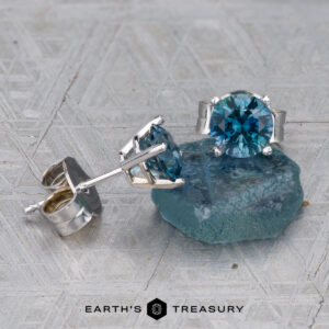 A pair of Montana Sapphire Earrings- Teal, 5.25mm (Heated) stud earrings rests on a rough blue stone, with Earth's Treasury logo at the bottom.