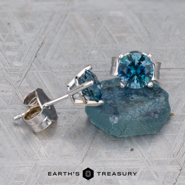 Montana Sapphire Earrings- Teal, 5.25mm (Heated) A pair of Montana Sapphire Earrings- Teal, 5.25mm (Heated) stud earrings rests on a rough blue stone, with Earth's Treasury logo at the bottom.