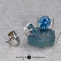A pair of Montana Sapphire Earrings in teal blue (5.25mm, heated) are displayed next to a rough blue gem on a textured surface, with the “Earth’s Treasury” logo at the bottom.