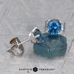 A pair of Montana Sapphire Earrings in teal blue (5.25mm, heated) are displayed next to a rough blue gem on a textured surface, with the “Earth’s Treasury” logo at the bottom.