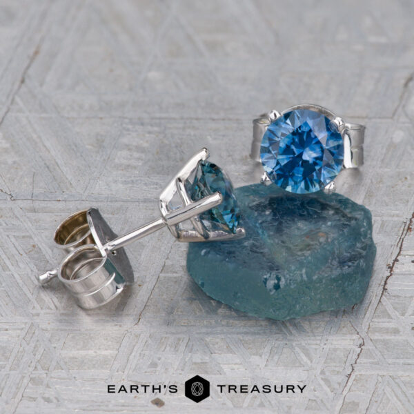 Montana Sapphire Earrings- TealBlue, 5.25mm (Heated) A pair of Montana Sapphire Earrings in teal blue (5.25mm, heated) are displayed next to a rough blue gem on a textured surface, with the “Earth’s Treasury” logo at the bottom.