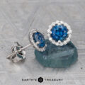 Montana Sapphire Diamond Halo Earrings (Rich Blue, 5.0 mm, Heated) with round sapphires encircled by sparkling diamonds, displayed on a textured surface featuring the "Earth's Treasury" logo.