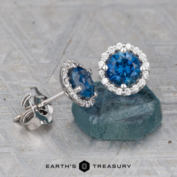 Montana Sapphire Diamond Halo Earrings, Rich Blue 5.0 mm (Heated Montana Sapphire Diamond Halo Earrings (Rich Blue, 5.0 mm, Heated) with round sapphires encircled by sparkling diamonds, displayed on a textured surface featuring the "Earth's Treasury" logo.