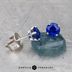 A pair of Deep Blue Sapphire Earrings, 5.0mm (Heated), in silver settings is shown with one earring resting on a rough blue stone atop a textured gray surface.