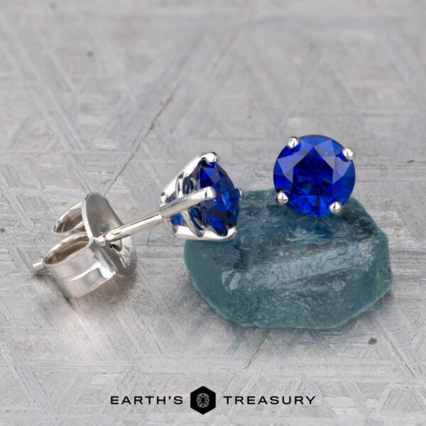 Deep Blue Sapphire Earrings, 5.0mm (Heated) A pair of Deep Blue Sapphire Earrings, 5.0mm (Heated), in silver settings is shown with one earring resting on a rough blue stone atop a textured gray surface.