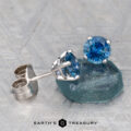 A pair of Montana Sapphire Earrings—medium blue, 5.25mm (heated), set in silver—displayed on a blue-green stone with the Earth’s Treasury logo below.