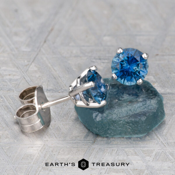 Montana Sapphire Earrings- Medium Blue, 5.25mm (Heated) A pair of Montana Sapphire Earrings—medium blue, 5.25mm (heated), set in silver—displayed on a blue-green stone with the Earth’s Treasury logo below.