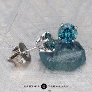 A pair of Montana Sapphire Earrings - Rich Teal, 5.25mm (Heated), set in silver are displayed on a rough teal stone, with the Earth’s Treasury logo visible below.