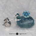 A pair of Montana Sapphire Earrings-Aqua, 5.25mm (Heated), featuring round-cut blue-green gemstones in silver settings, with one earring displayed on a rough sapphire piece.