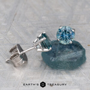 A pair of Montana Sapphire Earrings-Aqua, 5.25mm (Heated), featuring round-cut blue-green gemstones in silver settings, with one earring displayed on a rough sapphire piece.