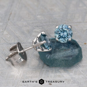 A pair of 5.25mm light aqua Montana sapphire stud earrings (heated) set in silver, displayed on a rough blue-green stone with the "Earth's Treasury" logo at the bottom.
