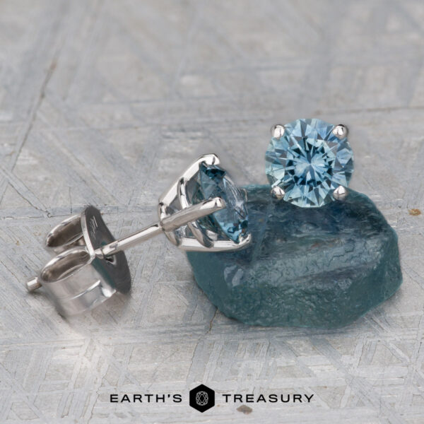 Montana Sapphire Earrings- Light Aqua, 5.25mm (Heated) A pair of 5.25mm light aqua Montana sapphire stud earrings (heated) set in silver, displayed on a rough blue-green stone with the "Earth's Treasury" logo at the bottom.