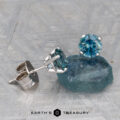Montana Sapphire Earrings- Teal, 5.25mm (Heated) are shown in silver settings, with one earring placed on a rough blue stone and both displayed on a textured gray surface.