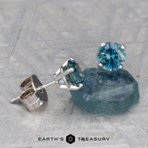 Montana Sapphire Earrings- Teal, 5.25mm (Heated) are shown in silver settings, with one earring placed on a rough blue stone and both displayed on a textured gray surface.