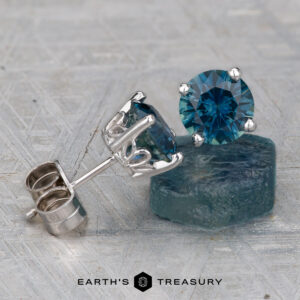 Montana Sapphire Earrings – Rich Teal, 6.2mm (Heated): A round blue sapphire stud in a silver setting beside a raw blue stone, on a textured surface with the Earth's Treasury logo below.