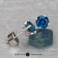 A pair of Montana Sapphire Earrings—rich blue, 6.4mm (heated)—in silver settings displayed on a textured surface, with one earring resting on a rough blue stone.