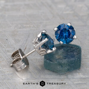 A pair of Montana Sapphire Earrings—rich blue, 6.4mm (heated)—in silver settings displayed on a textured surface, with one earring resting on a rough blue stone.