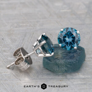 A pair of Montana Sapphire Earrings- Teal, 6.0mm (Heated) are shown in silver settings, with one earring resting on a rough blue stone and the Earth's Treasury logo displayed at the bottom.