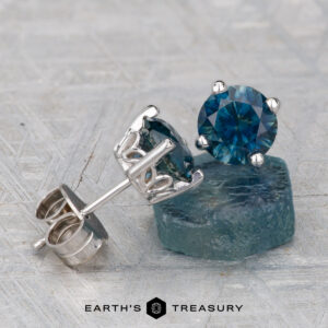 Montana Sapphire Earrings in deep peacock blue (6.5mm, heated) feature round gemstones in silver settings, displayed on a textured gray surface with one earring resting on a blue stone.