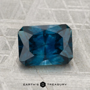A rectangular, deep blue 2.40-carat Montana Sapphire (Heated) with faceted edges is displayed on a patterned gray surface, with the text "EARTH'S TREASURY" at the bottom.
