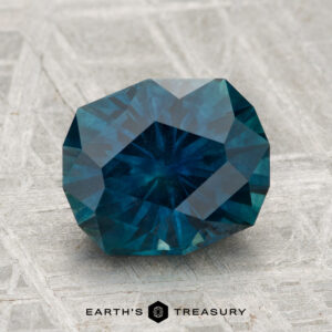 A 2.97-carat heated Montana sapphire with a deep blue hue is displayed on a textured gray surface, with "Earth's Treasury" shown below.
