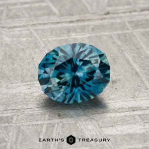 A 1.03-carat heated Montana Sapphire, round brilliant-cut and blue in color, is displayed on a textured gray surface with the Earth's Treasury logo at the bottom.