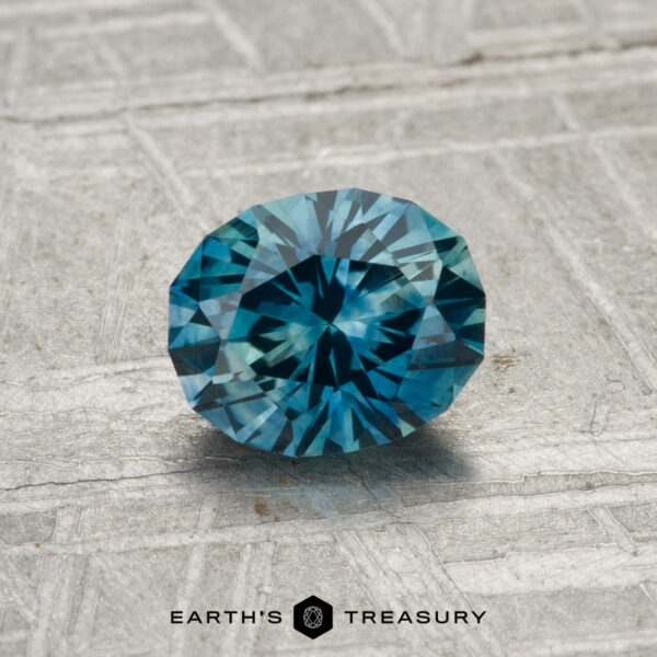 1.03-Carat Montana Sapphire (Heated) A 1.03-carat heated Montana Sapphire, round brilliant-cut and blue in color, is displayed on a textured gray surface with the Earth's Treasury logo at the bottom.